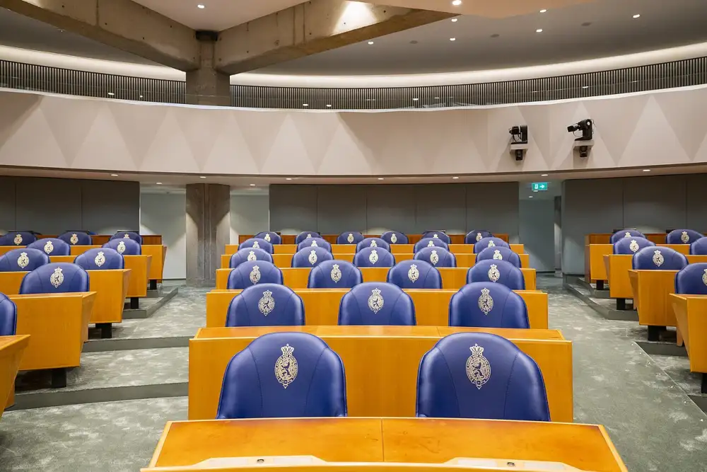 Lege plenaire zaal van de Tweede Kamer op de tijdelijke locatie, blauwe zetels met het Kamerwapen